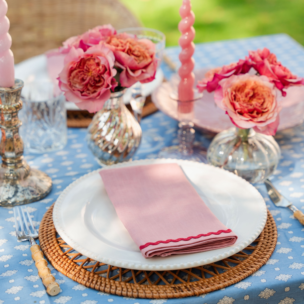 Table setting with pink napkin, flowers, and cutlery on a floral tablecloth.
