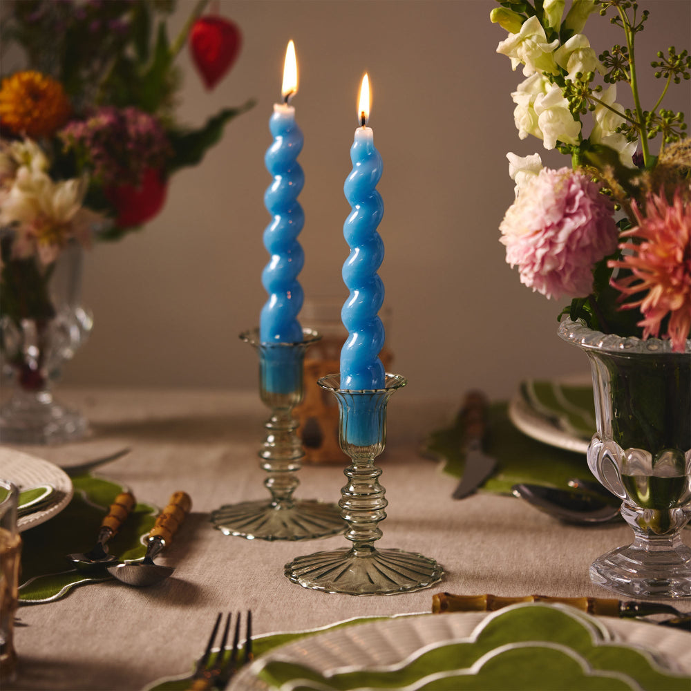 Two blue spiral candles in glass holders on a table with floral arrangements and cutlery.
