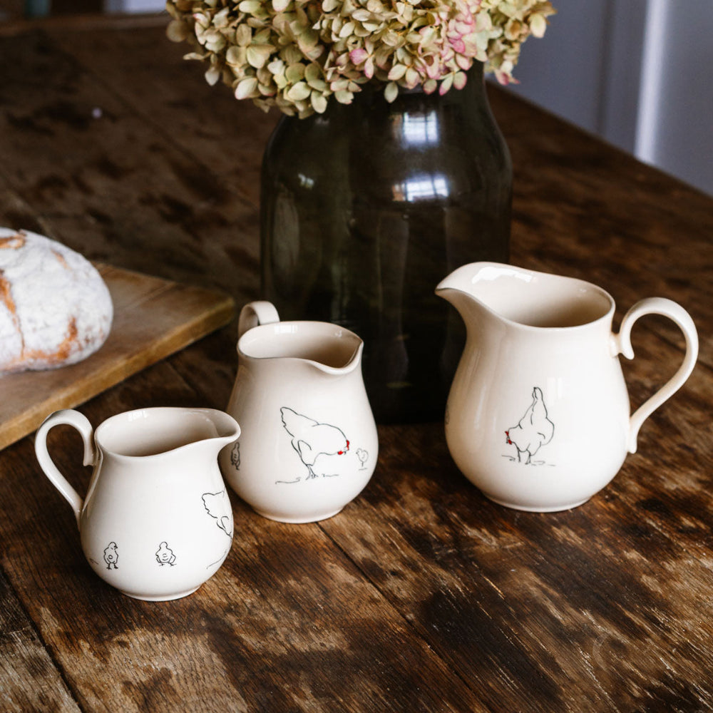 Three ceramic pitchers with chicken illustrations on a wooden table, accompanied by a vase with flowers.