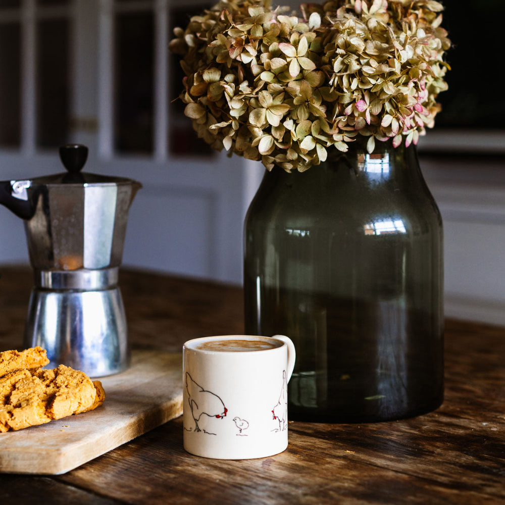 Café au lait in a white mug with red chicken designs, a green vase with dried flowers, and a silver espresso pot on a wooden table.