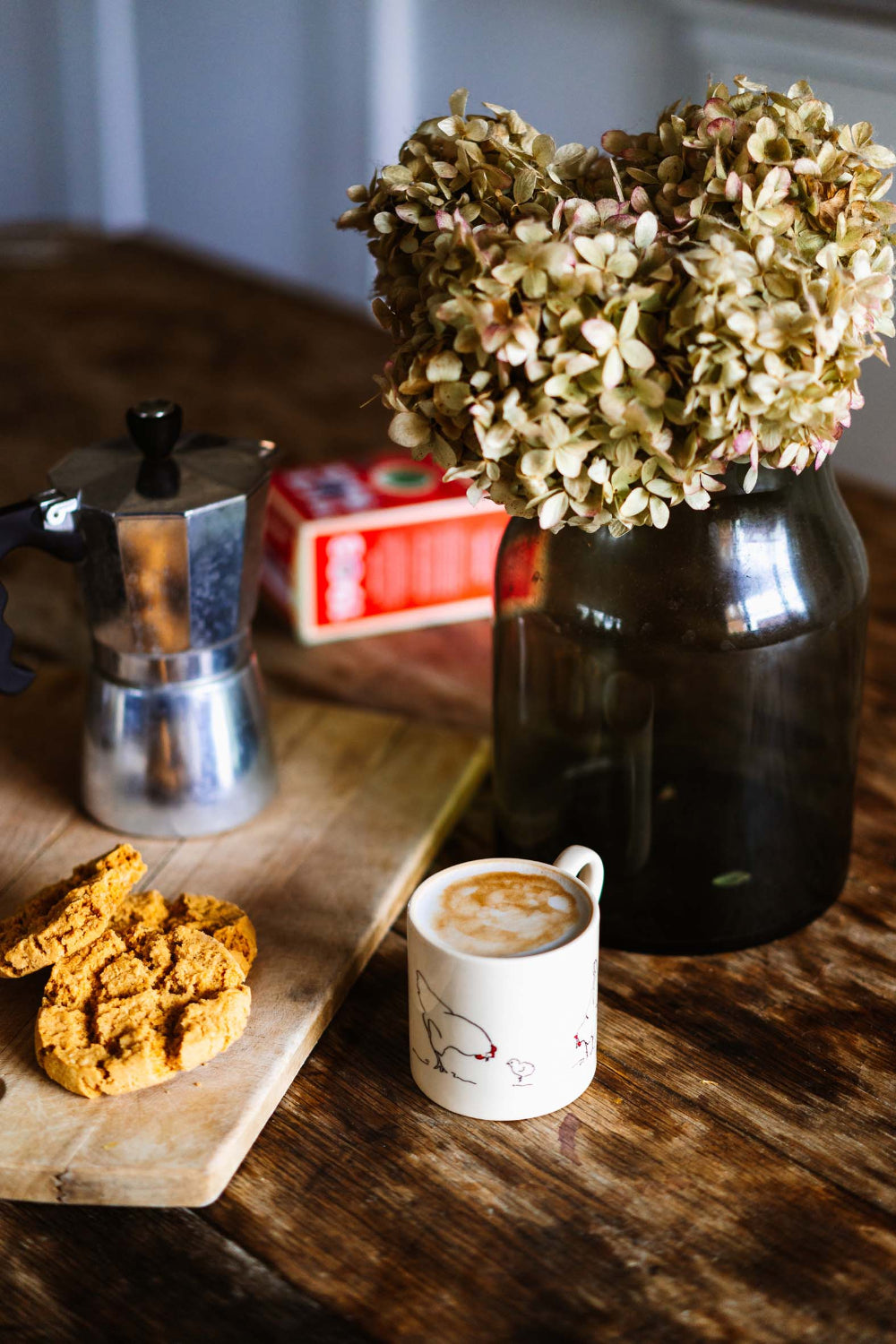Café au lait in a white cup with a design, cookies, and a coffee pot on a wooden table.