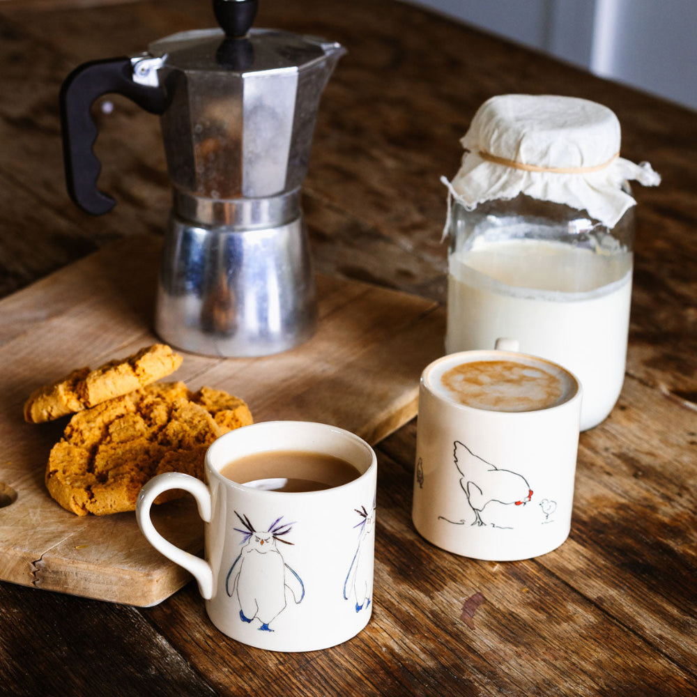 Two mugs with coffee and milk, a stovetop espresso maker, and cookies on a wooden surface.