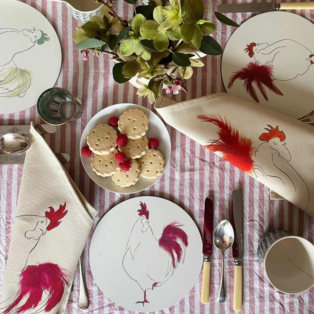 Table setting with rooster-themed plates, napkins, and cutlery on a red and white checkered tablecloth.