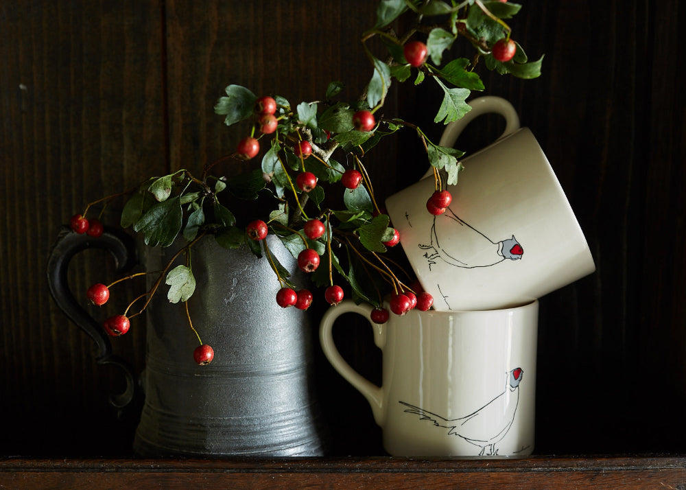 Two ceramic mugs with red berries and green leaves on a wooden surface.