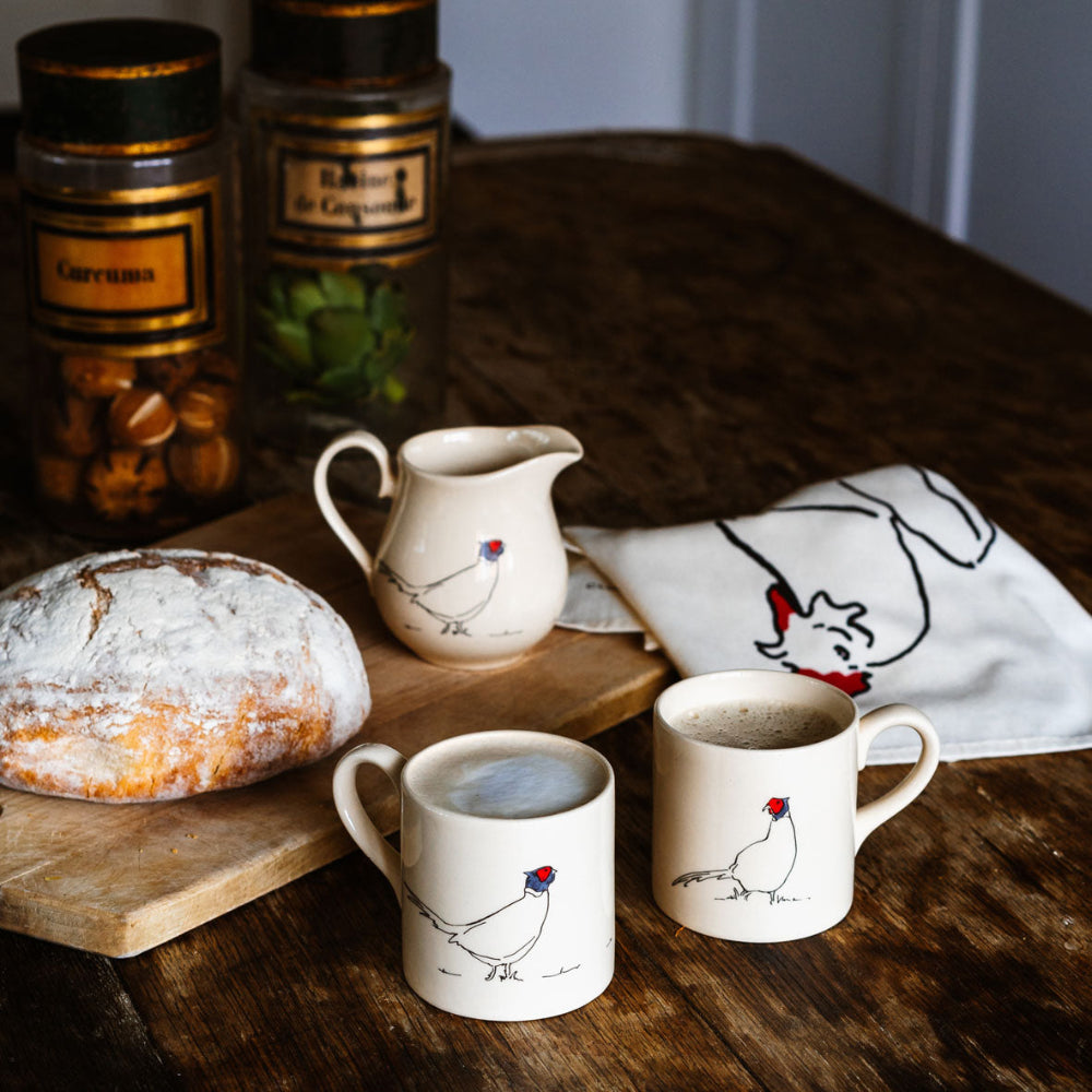 Two mugs with Pheasant designs on a wooden table with bread and a pitcher.