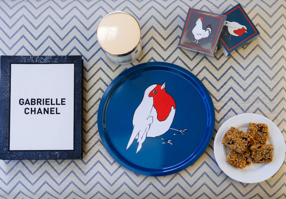 Blue tray with a red and white Robin design, a mug, a book titled 'Gabrielle Chanel', and a plate of cookies on a zigzag patterned surface.