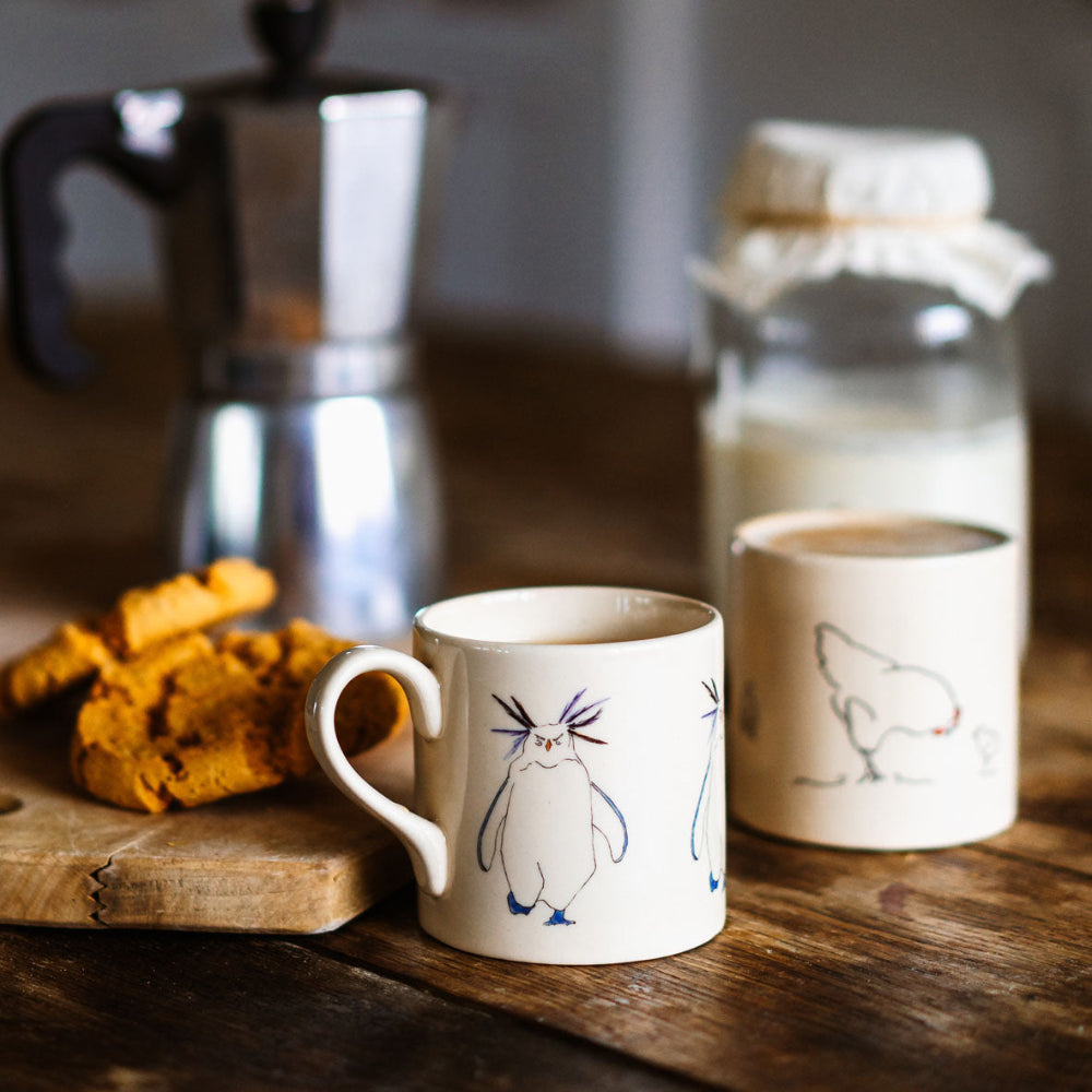 Two ceramic mugs with chicken and rockhopper illustrations on a wooden surface with cookies and a milk bottle in the background.