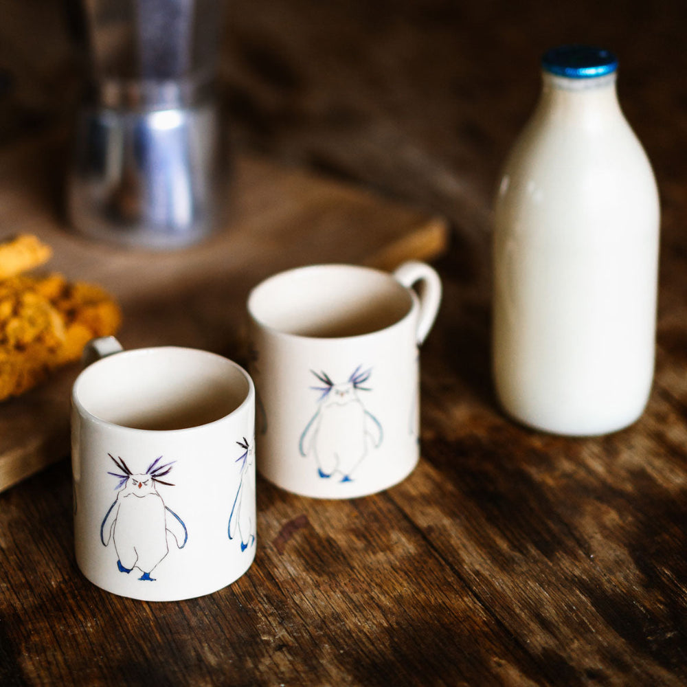 Two ceramic mugs with penguin designs on a wooden surface next to a bottle of milk.