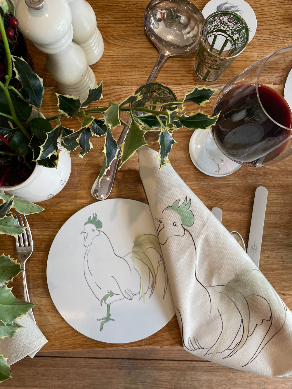 Table setting with a decorative plate and napkin featuring rooster designs, surrounded by holly leaves and a glass of red wine.