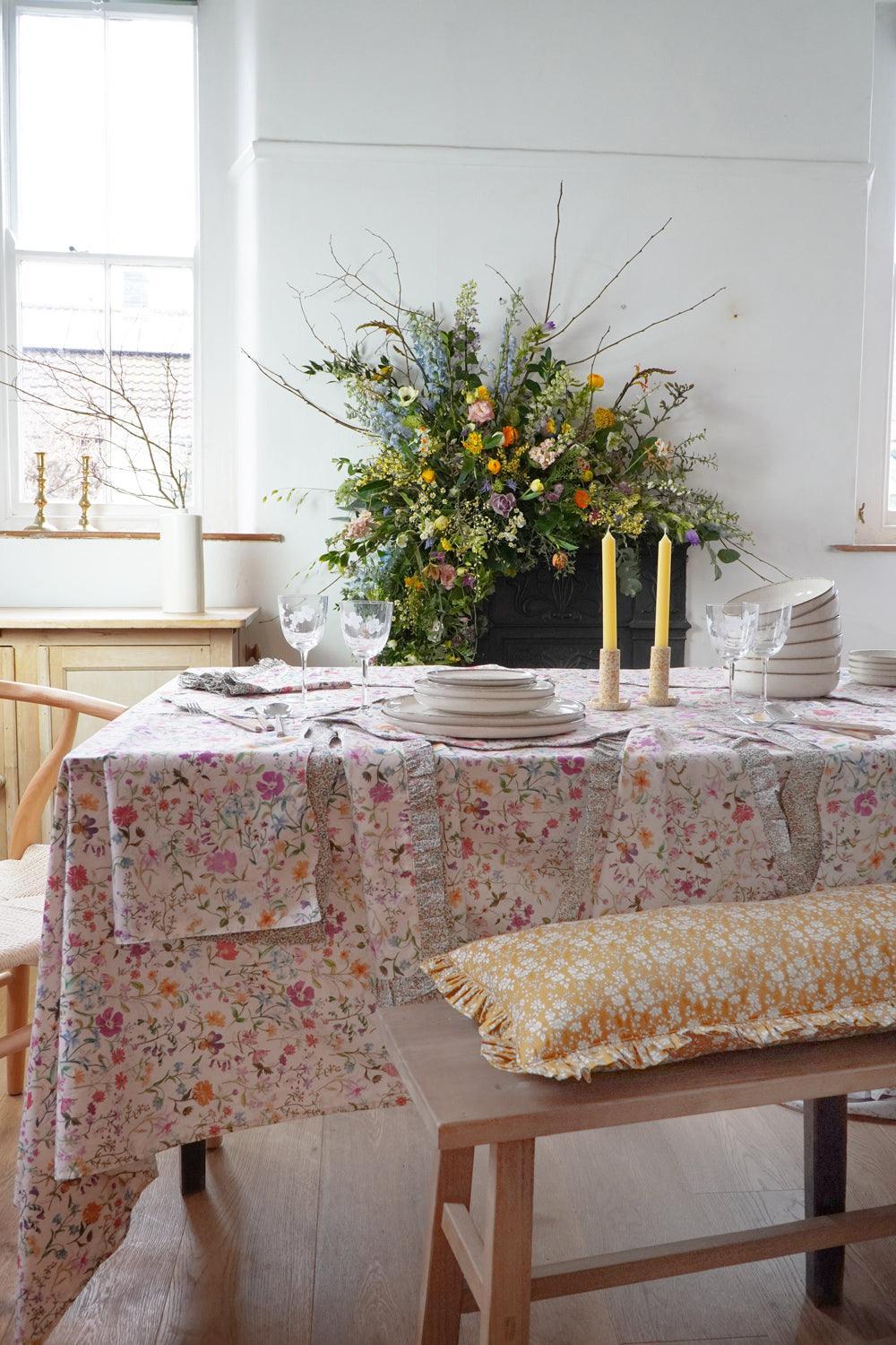 Dining table set with a floral tablecloth, plates, and candles in a bright room.