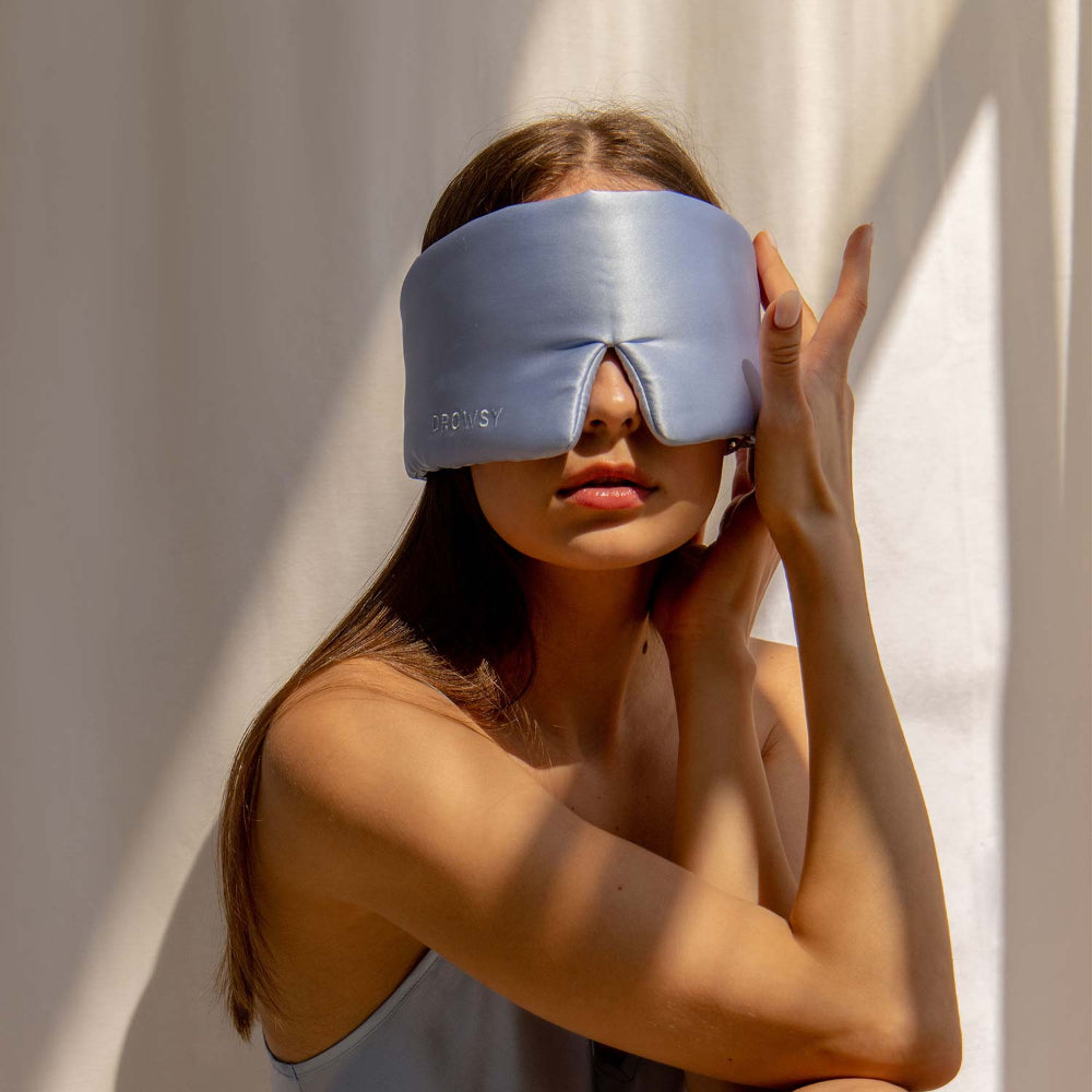 Person wearing a blue silk sleep mask against a neutral background