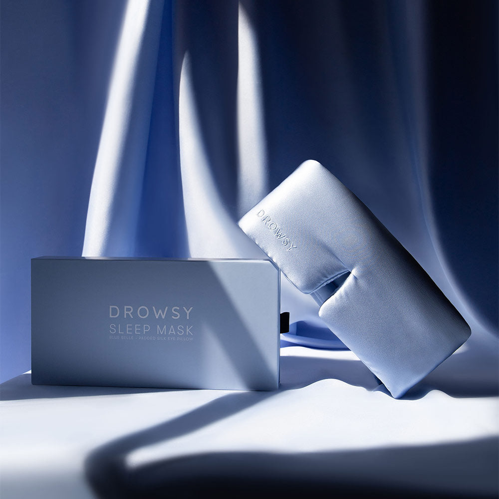 Drowsy Blue Belle Silk sleep mask with packaging in front of a silk draped fabric background