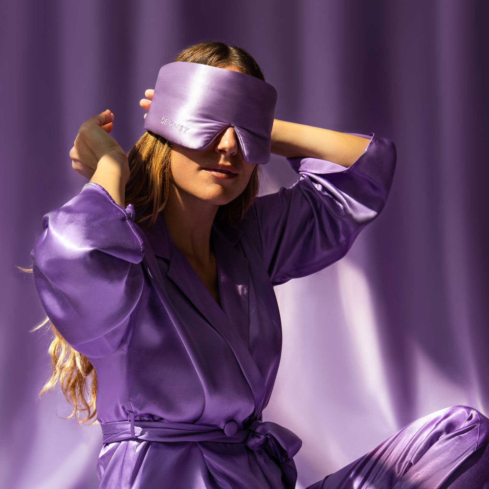 Person wearing a purple silk robe and eye mask against a purple curtain.