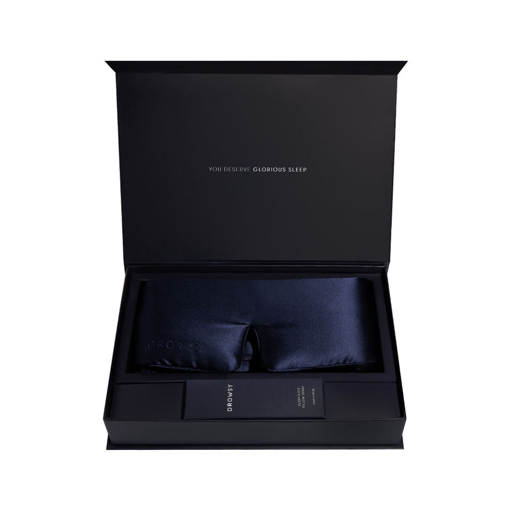 Open black box containing a navy blue silk sleep mask and pillow spray on a white background