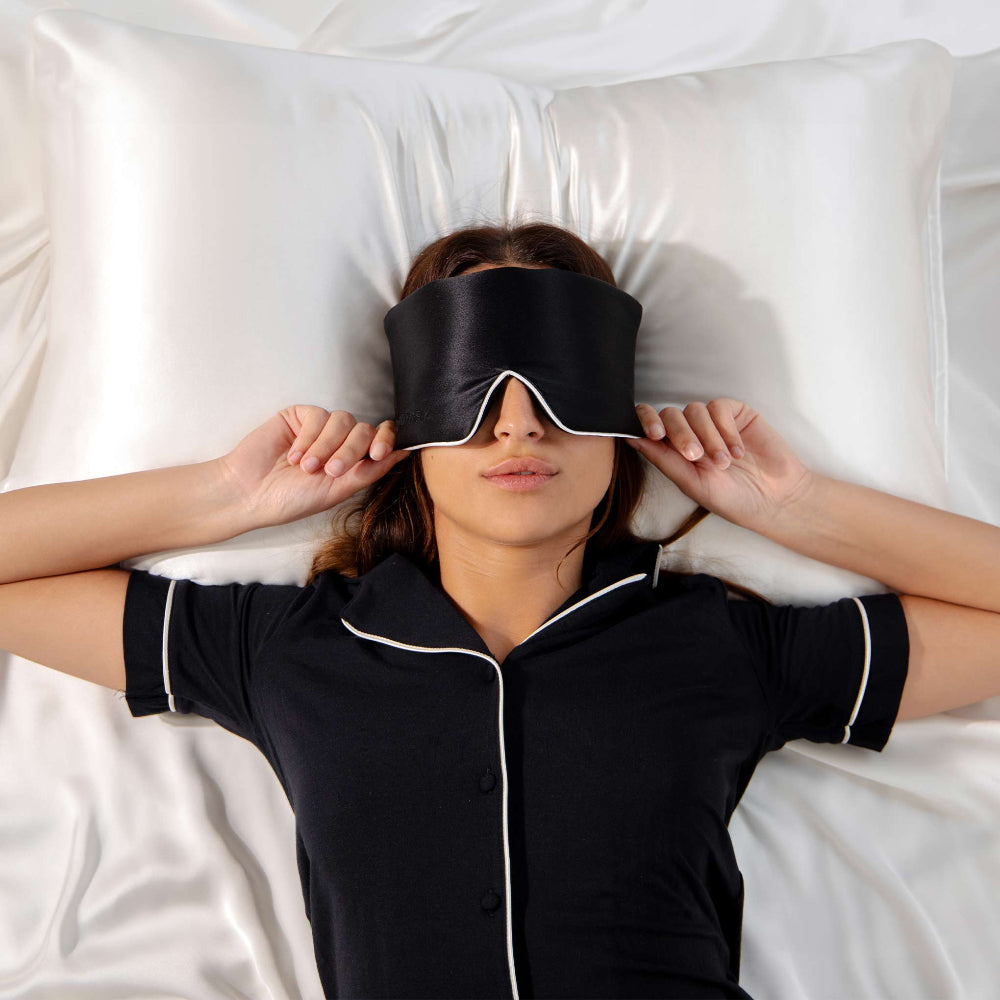 Woman laying on silk bedsheets in black pyjamas with white piping edge holding onto her black silk sleep mask with white edge piping.