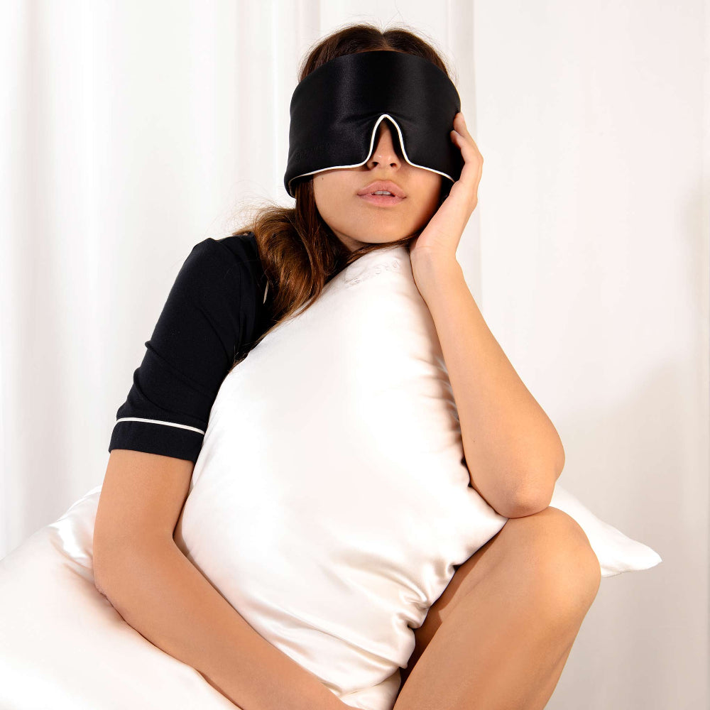 Woman holding onto a white silk pillowcase with black silk sleep mask with white piping edge