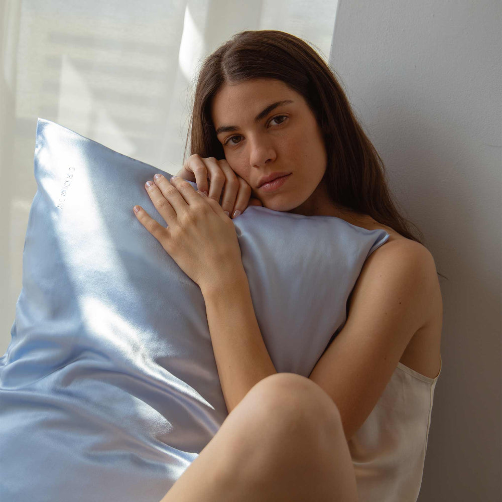 Woman leaning her head and holding a Drowsy Blue Belle Silk pillow in a light bedroom setting