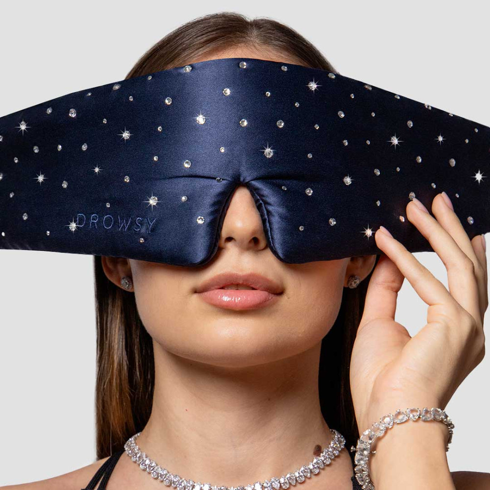 Person wearing a navy blue sleep mask with silver crystals on a plain background