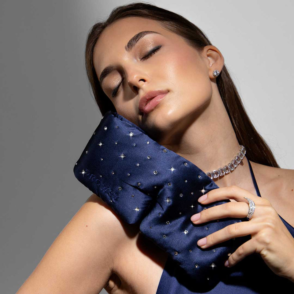 Woman holding a blue silk sleep mask with crystals near shoulder against a grey background