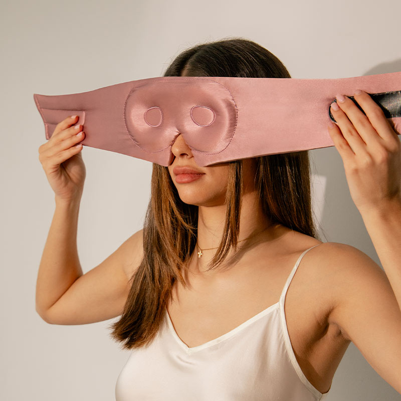 A person holding up a pink silk eyelash protecting mask, showing the interior contour for the eyes.