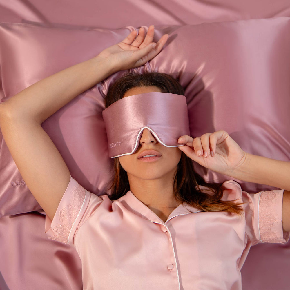 Person wearing a pink satin sleep mask and pajamas, lying in pink bedding.