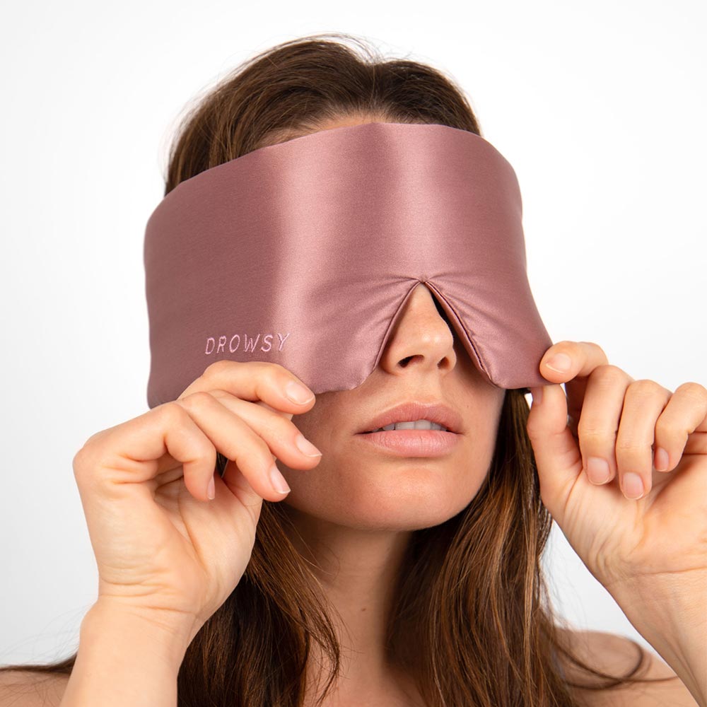 Woman holding over her eyes a Drowsy Damask Rose pink sleep mask on a white background