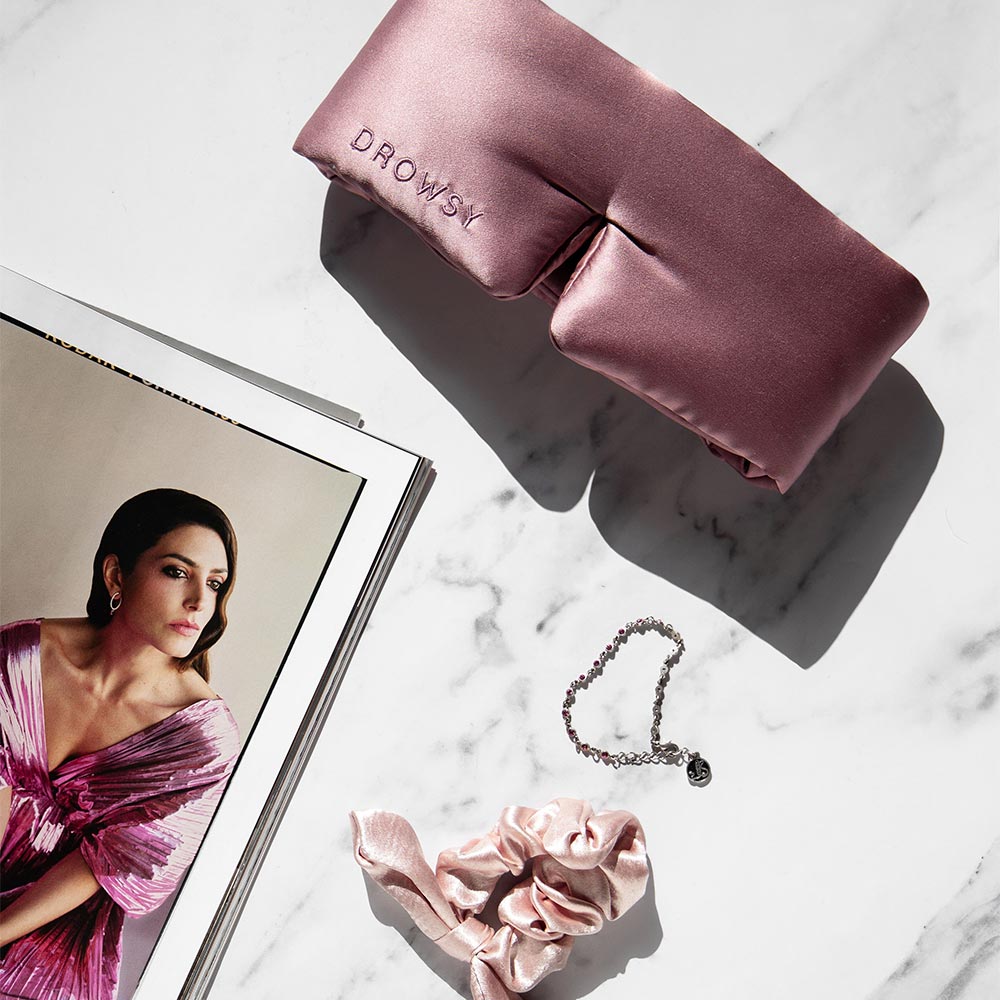 Drowsy Damask Rose Pink Silk sleep mask, pink scrunchie, and necklace on a marble surface with a magazine.