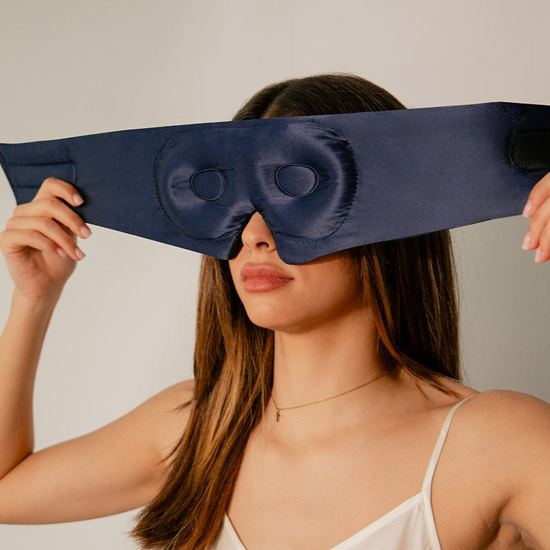 A person is holding up a midnight blue silk sleep mask covering their eyes, showcasing the product's design and fit.