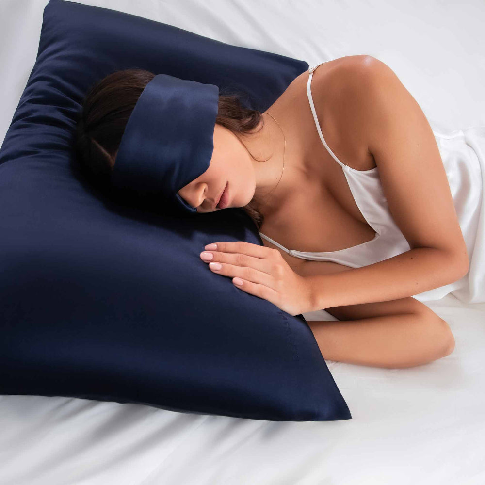 Woman laying down on white bedding with a midnight blue silk pillowcase and matching sleep mask