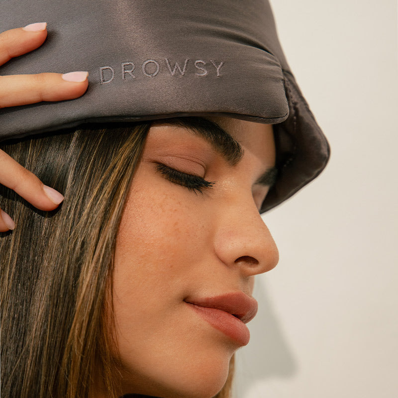 Closeup image of woman wearing a grey silk sleep eye mask with 'DROWSY' text.
