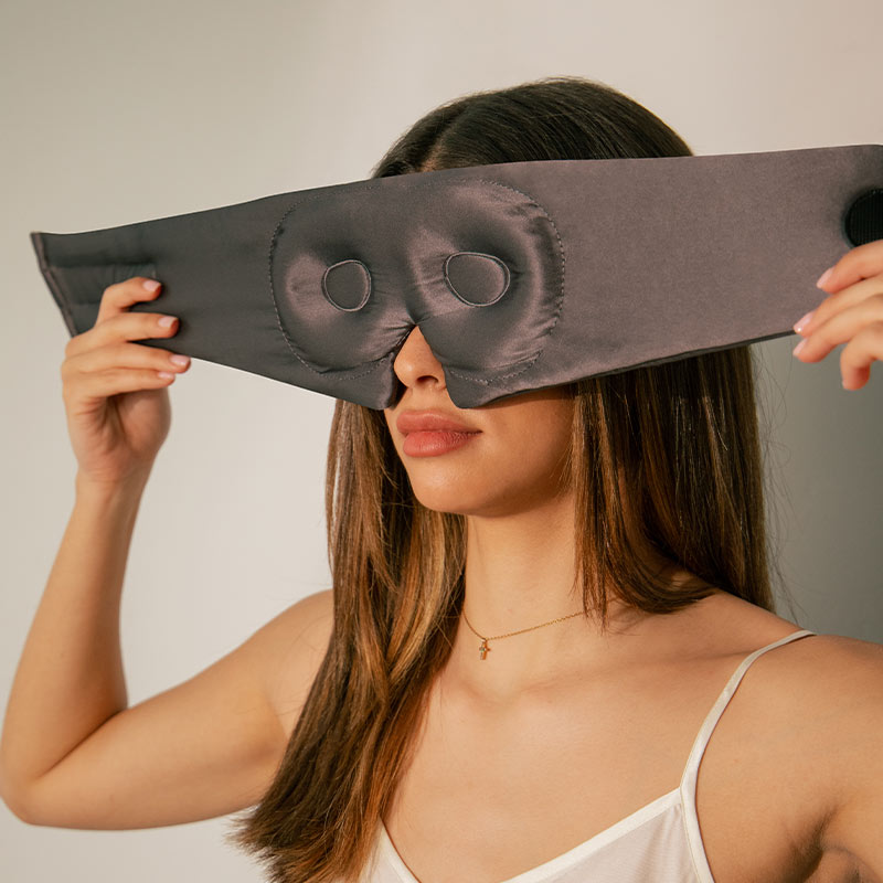 A person is holding up a grey silk sleep mask in front of their face, showing the contoured eyecups and the soft texture of the material.