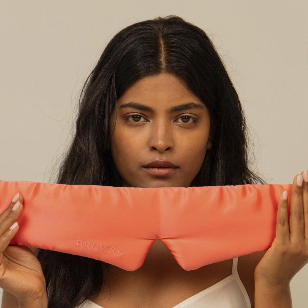 Woman holding a coral silk sleep mask slightly underneath chin.