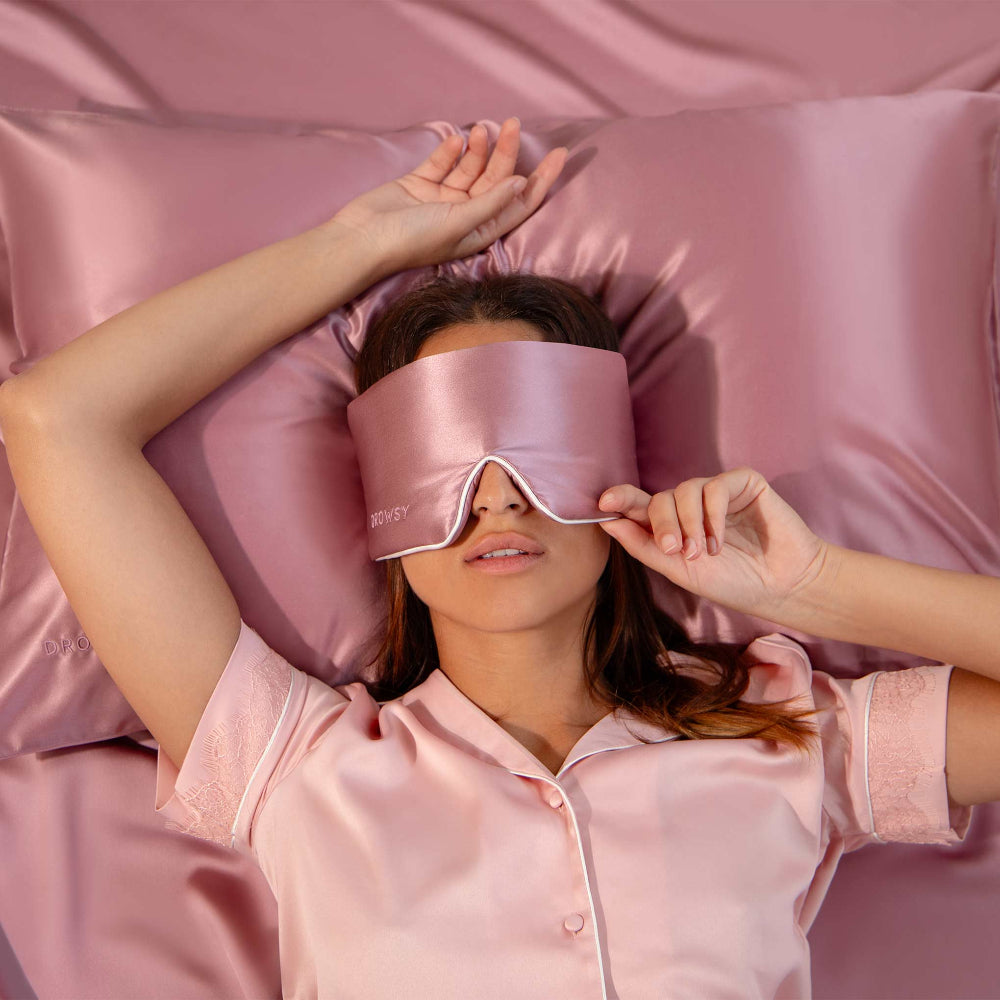 Woman laying on a bed wearing a pink silk sleep mask and matchin silk pillows.