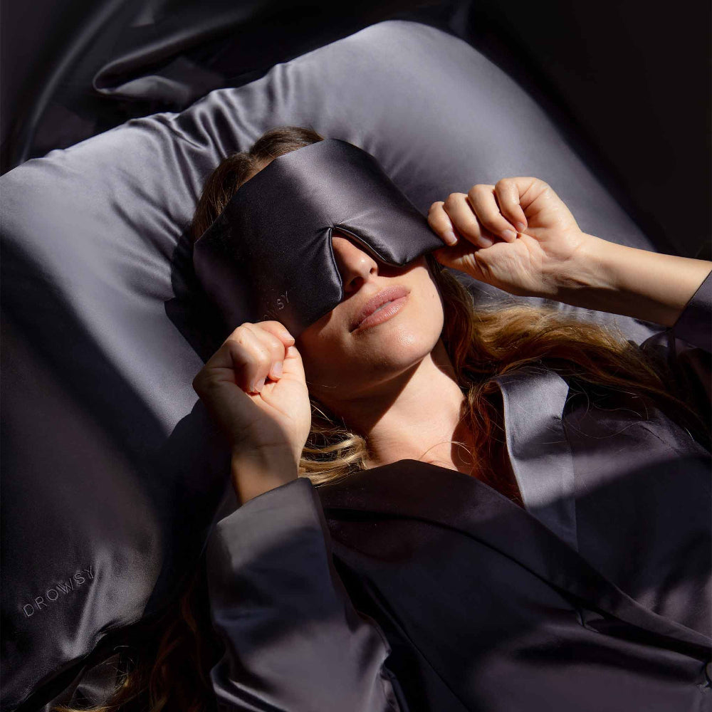 Woman laying down holding a moonlight shadow eye mask to face laying on moonlight shadow silk pillowcase
