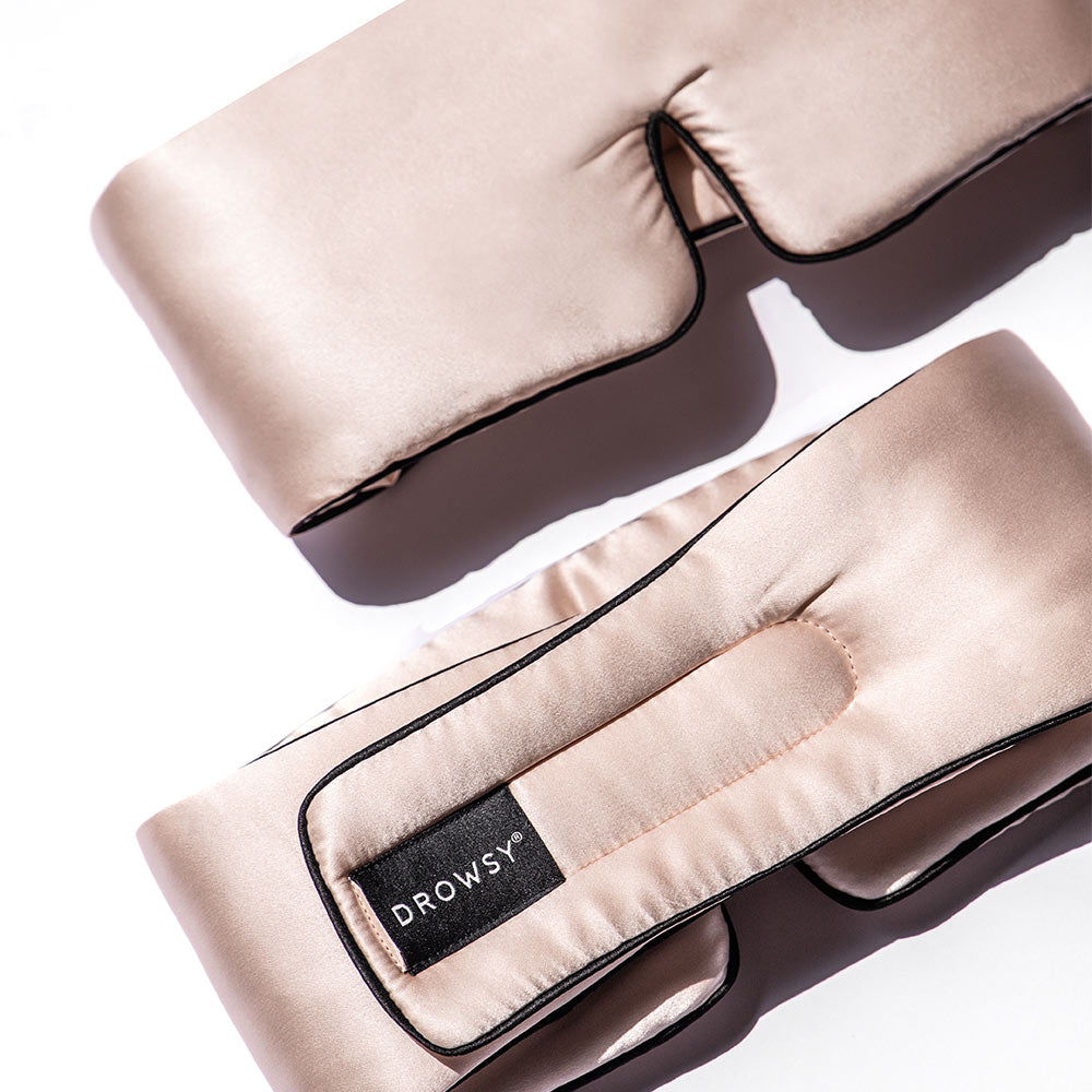 A pastel pink silk sleep mask with black piping and a Drowsy brand tag, laying flat against a light background.