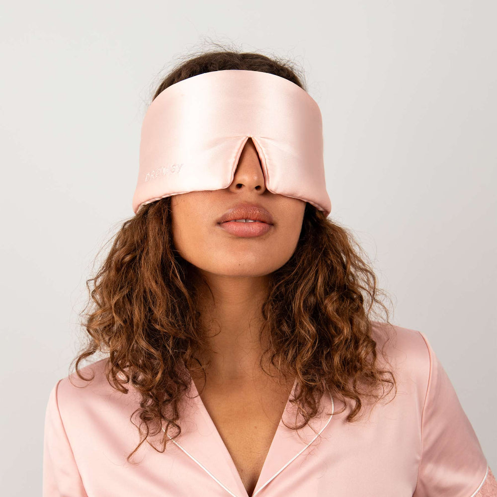 Woman with brown curly hair wearing a pink silk sleep mask around her eyes and pink silk pyjamas against a plain background.