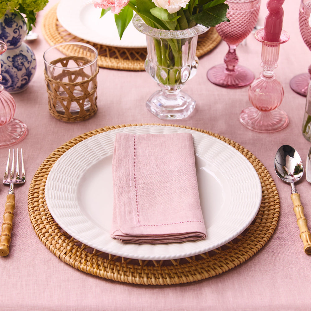 Table setting with a white plate, pink napkin, and wicker placemat on a pink tablecloth.
