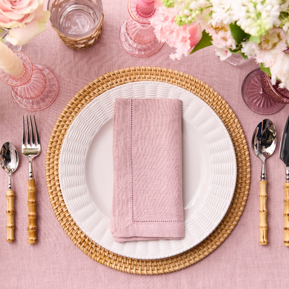 Table setting with pink napkin, white plate, and wicker charger on a pink tablecloth.