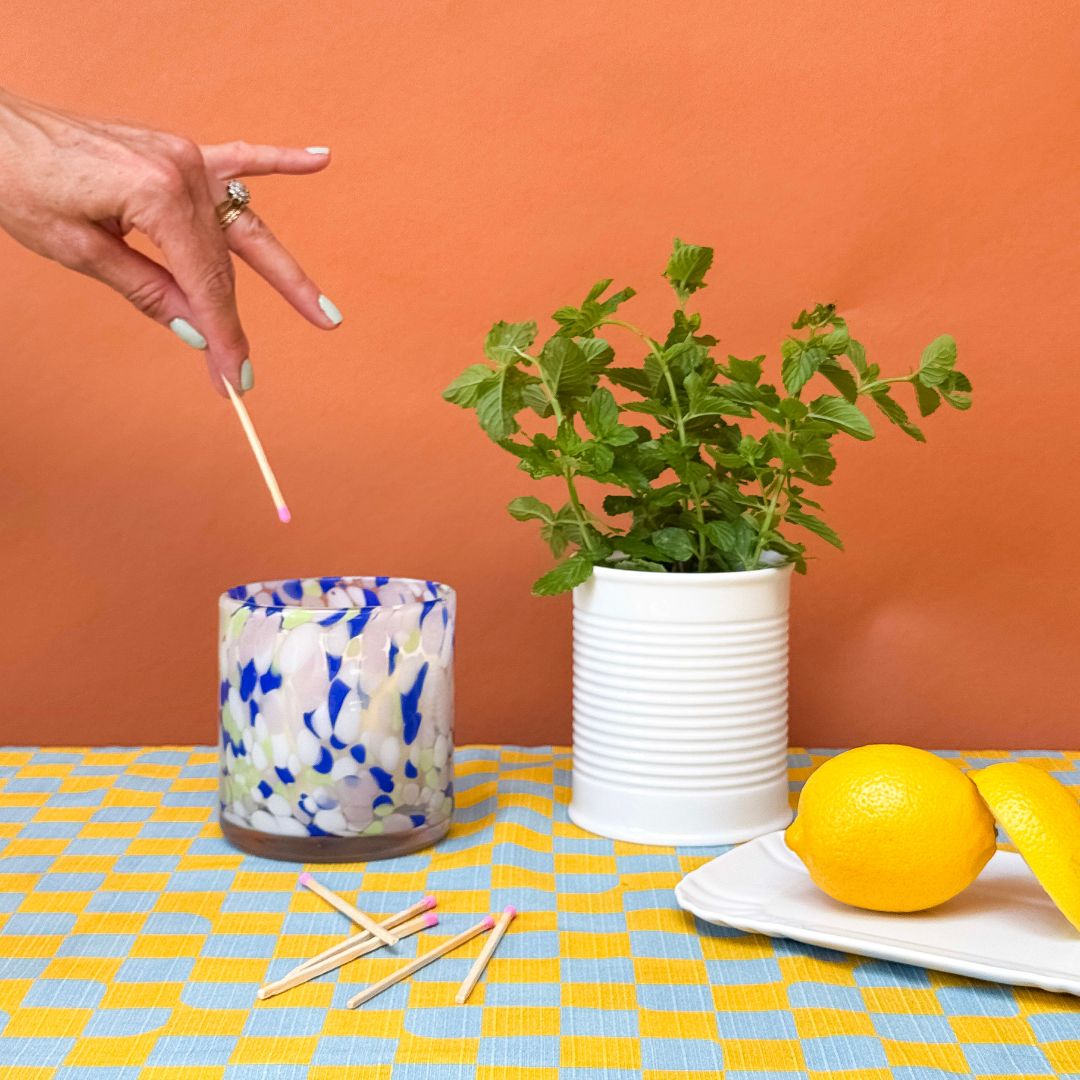 Hand holding a match near a colourful candle, plant, and lemons on a checkered tablecloth.
