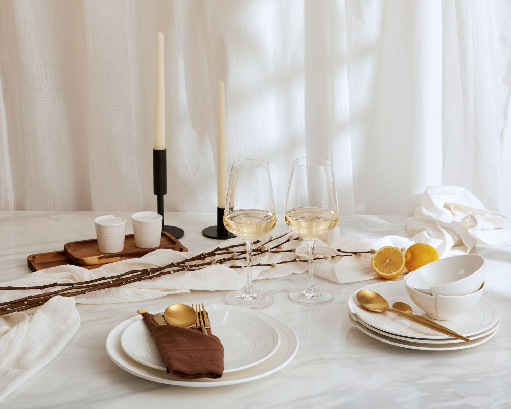 Elegant table setting with white plates, gold cutlery, and candles against a white curtain background.