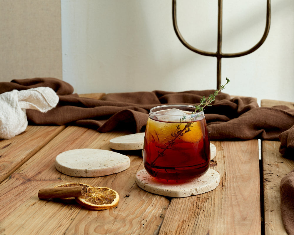 Glass of iced tea with a sprig of rosemary on a wooden table with a brown cloth and dried oranges on the Elliot Travertine Coasters.