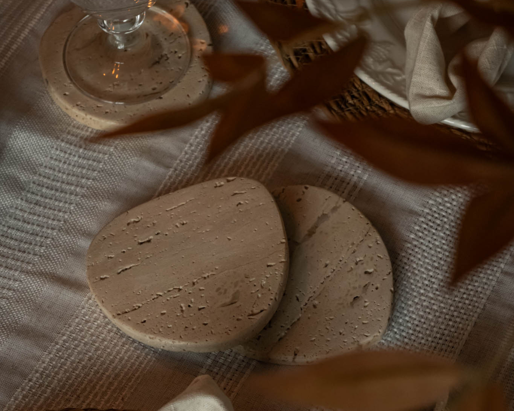 Two travertine coasters on a textured fabric surface with a glass and basket in the background.