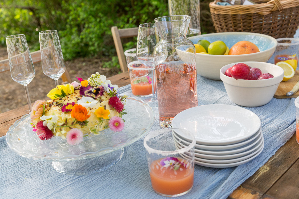 Outdoor table setting with a floral cake, drinks, and fruit on a blue tablecloth.