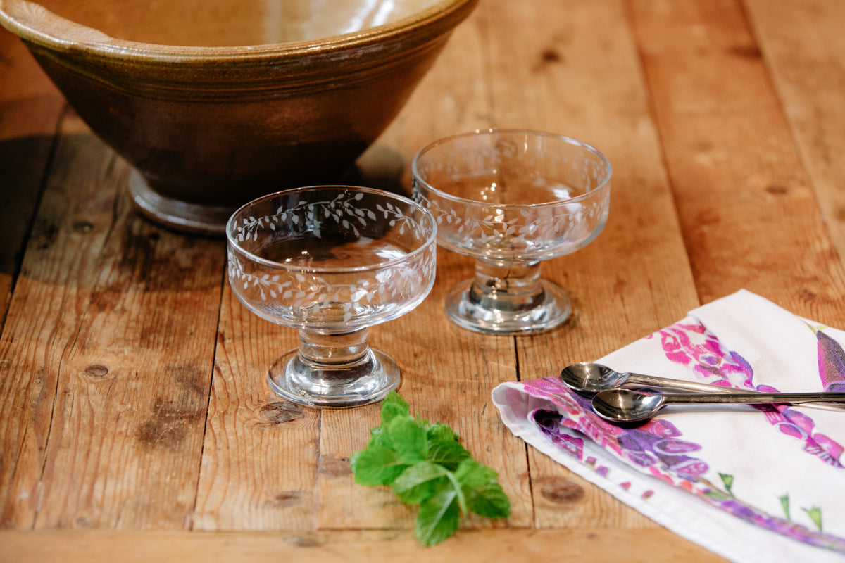 Two glass dessert dishes on a wooden table with a floral napkin and spoon.