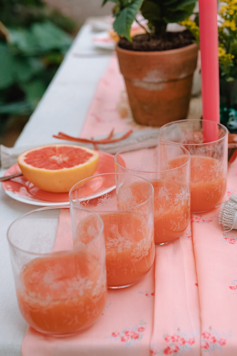 Orange juice in glasses on a floral tablecloth with a pink candle and potted plant in the background.