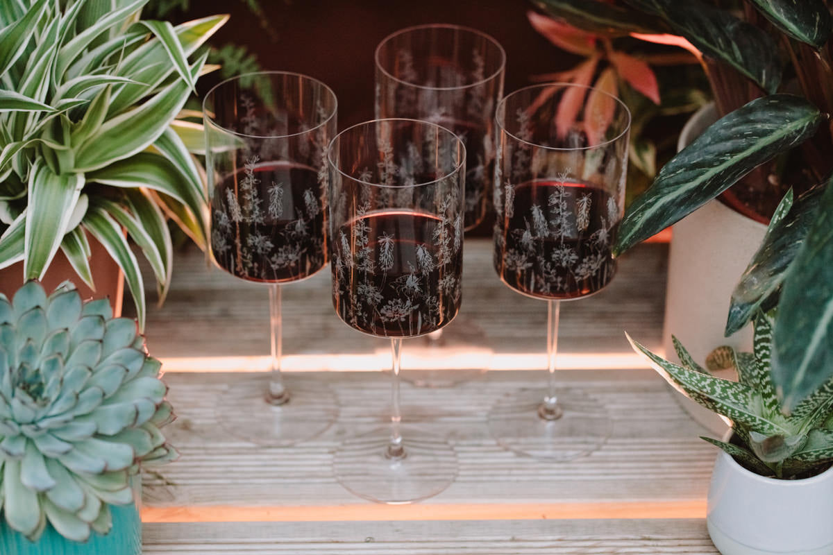 Four wine glasses with red wine on a wooden surface surrounded by plants