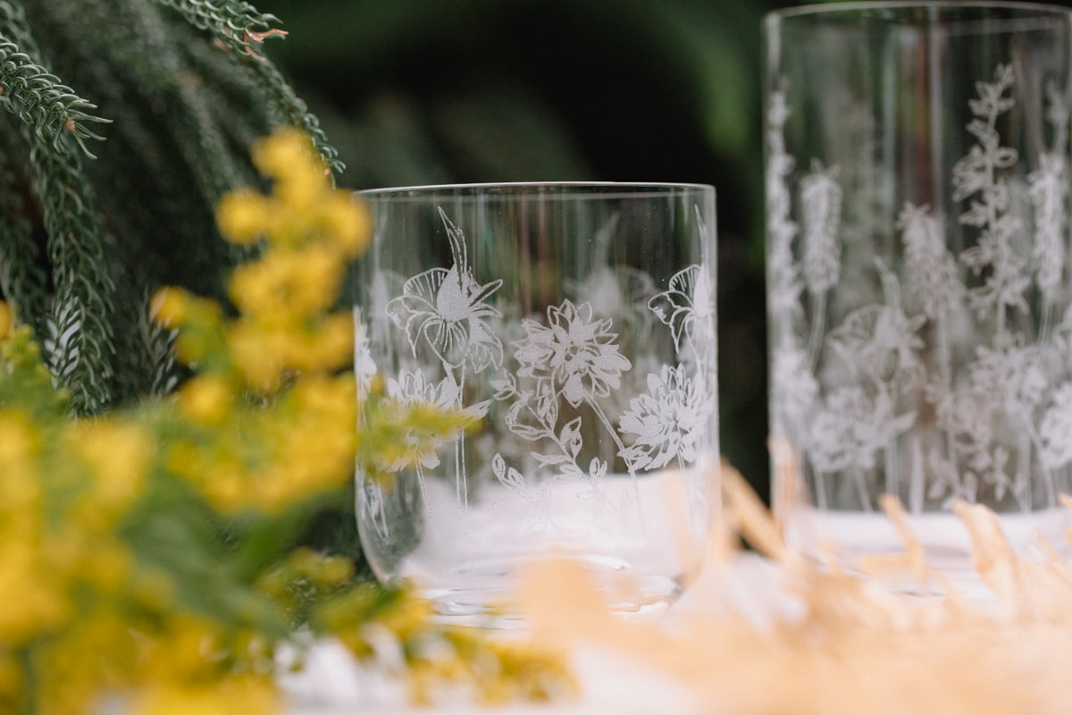 Two etched glass tumblers with floral designs on a blurred natural background