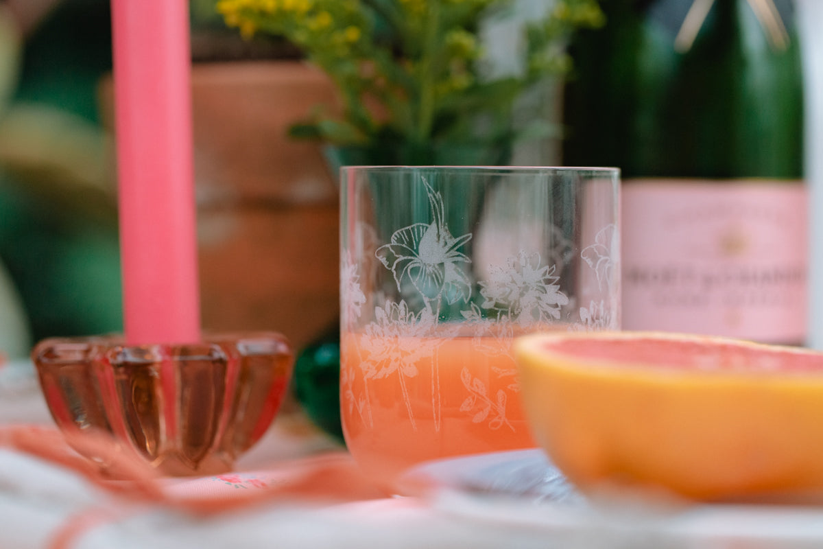 Glass with floral design filled with a pink drink, surrounded by orange slices and a pink candle.