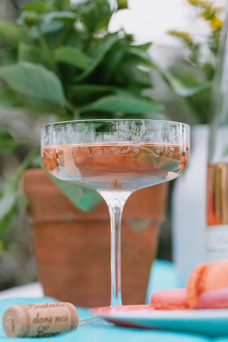 Cocktail glass with a drink on a table outdoors, with plants and a bottle in the background.