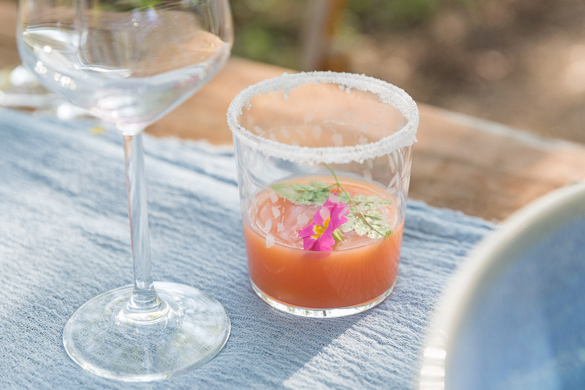 Small glass with a pink drink and a flower on a blue tablecloth, next to a wine glass.